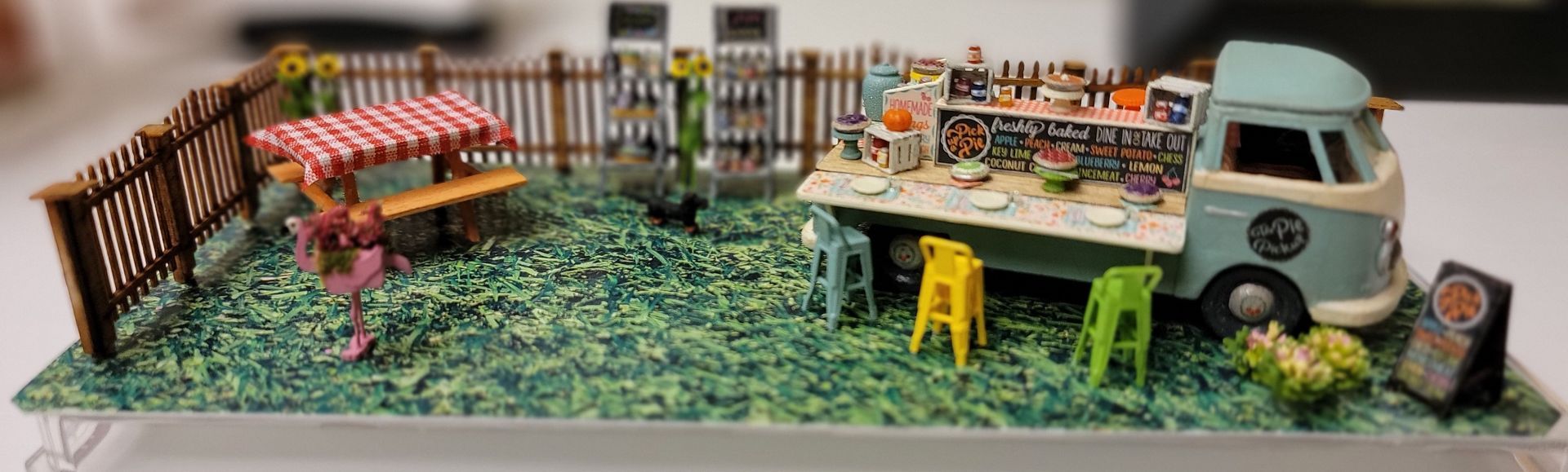 Miniature outdoor café scene with a red-and-white canopy, yellow stools, and a food truck on grass.