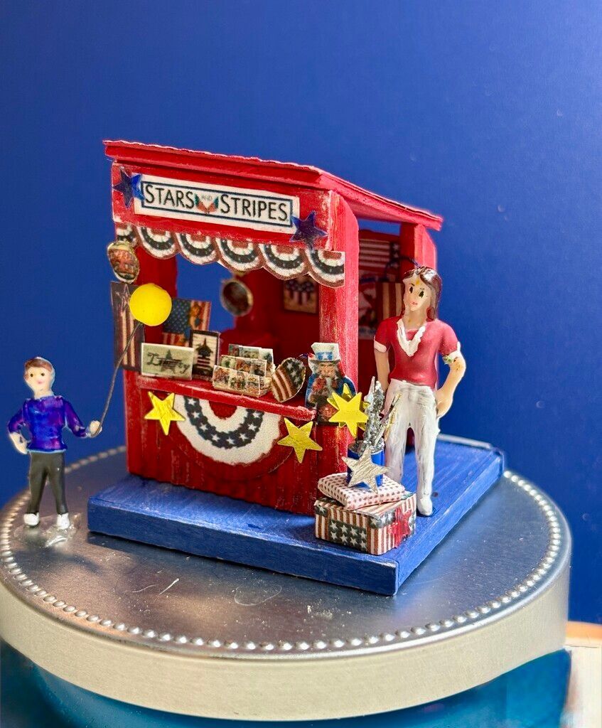 Miniature toy cottage with blue roof, lit storefront, and small figures on a wooden base.
