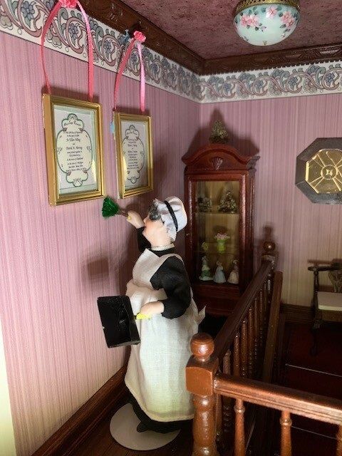 Miniature Victorian dollhouse hallway with a maid figurine holding a book beside framed wall art.