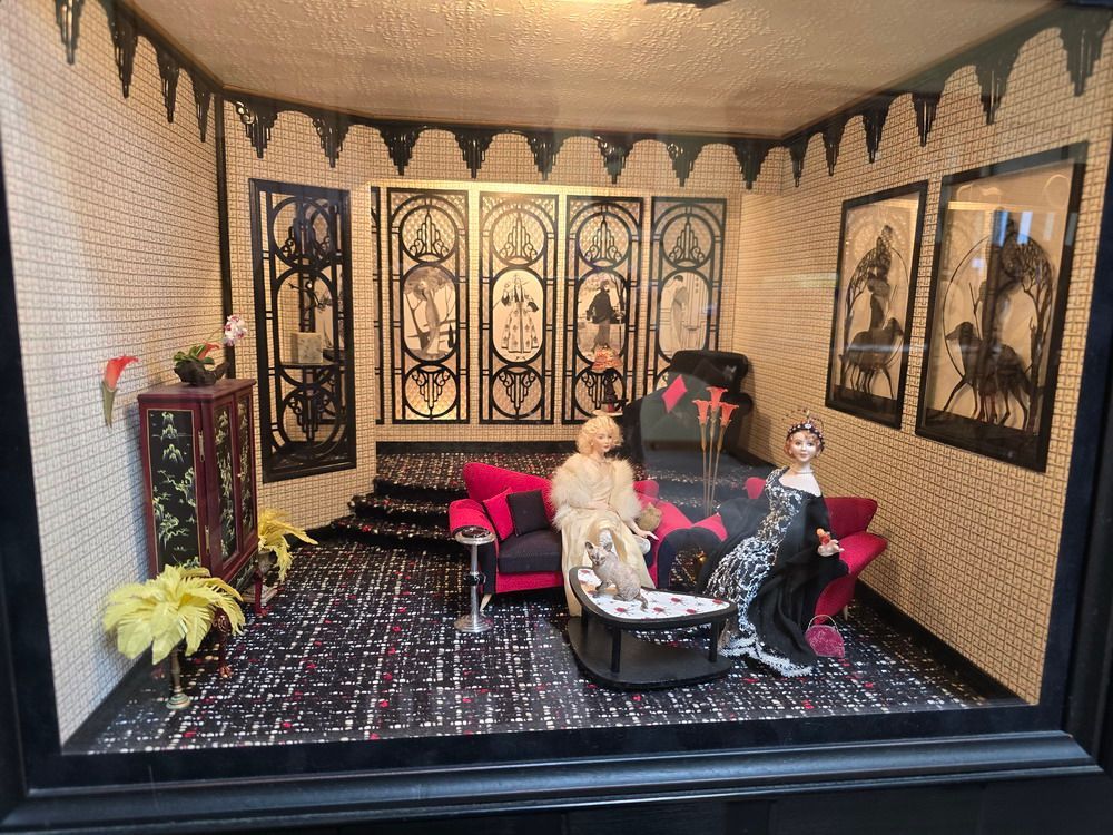 Miniature vintage-style living room with two seated dolls, black-and-red furniture, and ornate wall decor