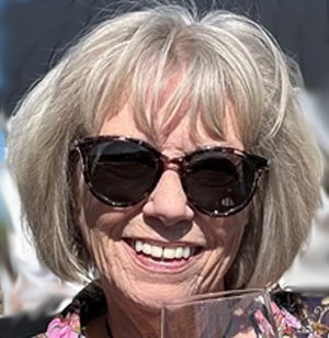 Smiling woman with short gray hair and black sunglasses holding a clear glass outdoors