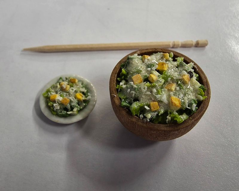 Miniature bowl of rice topped with vegetables beside a tiny plate and chopstick on a gray surface