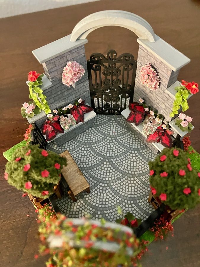 Miniature courtyard with arched gate, patterned patio, and pink flowers in a landscaped garden scene