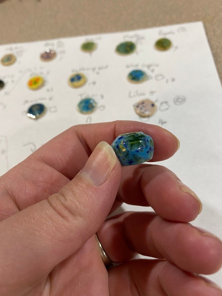 Hand holding a small blue-green glass bead in front of a bead color chart