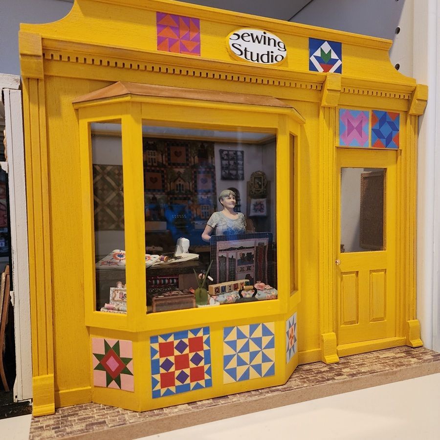 Bright yellow sewing studio storefront with quilt-patterned window panels and a mannequin inside.