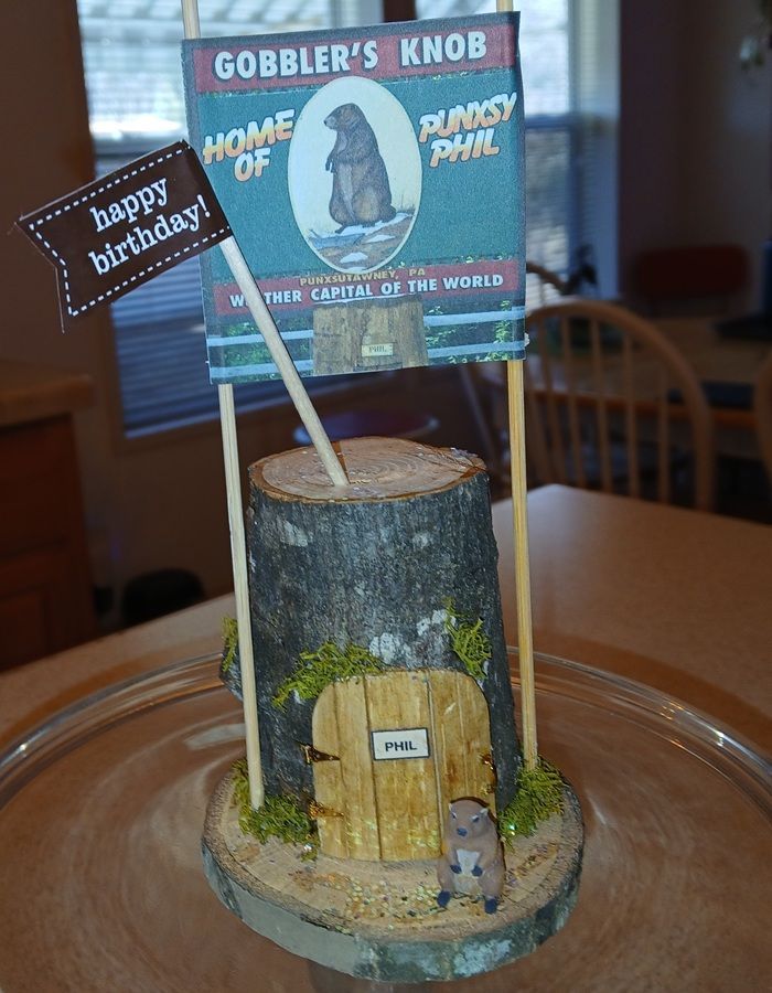 Birthday cake decorated like a wooden tree stump with a small sign and a “Happy Birthday” flag on a plate