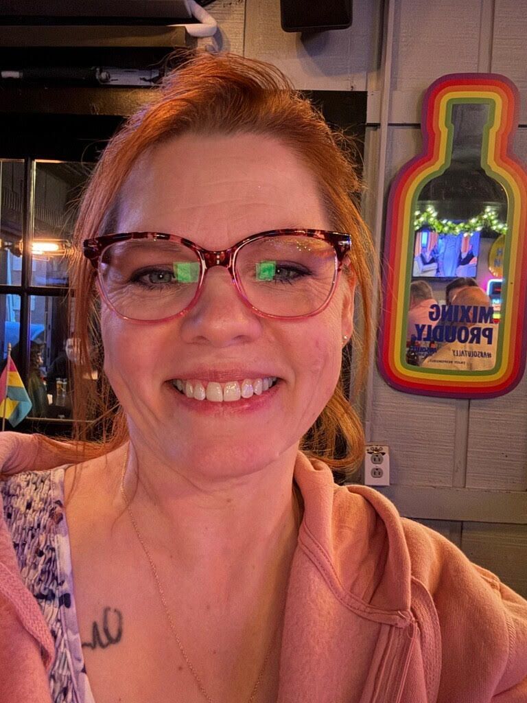 Smiling woman with glasses in a pink hoodie, seated indoors beside a colorful neon bottle sign