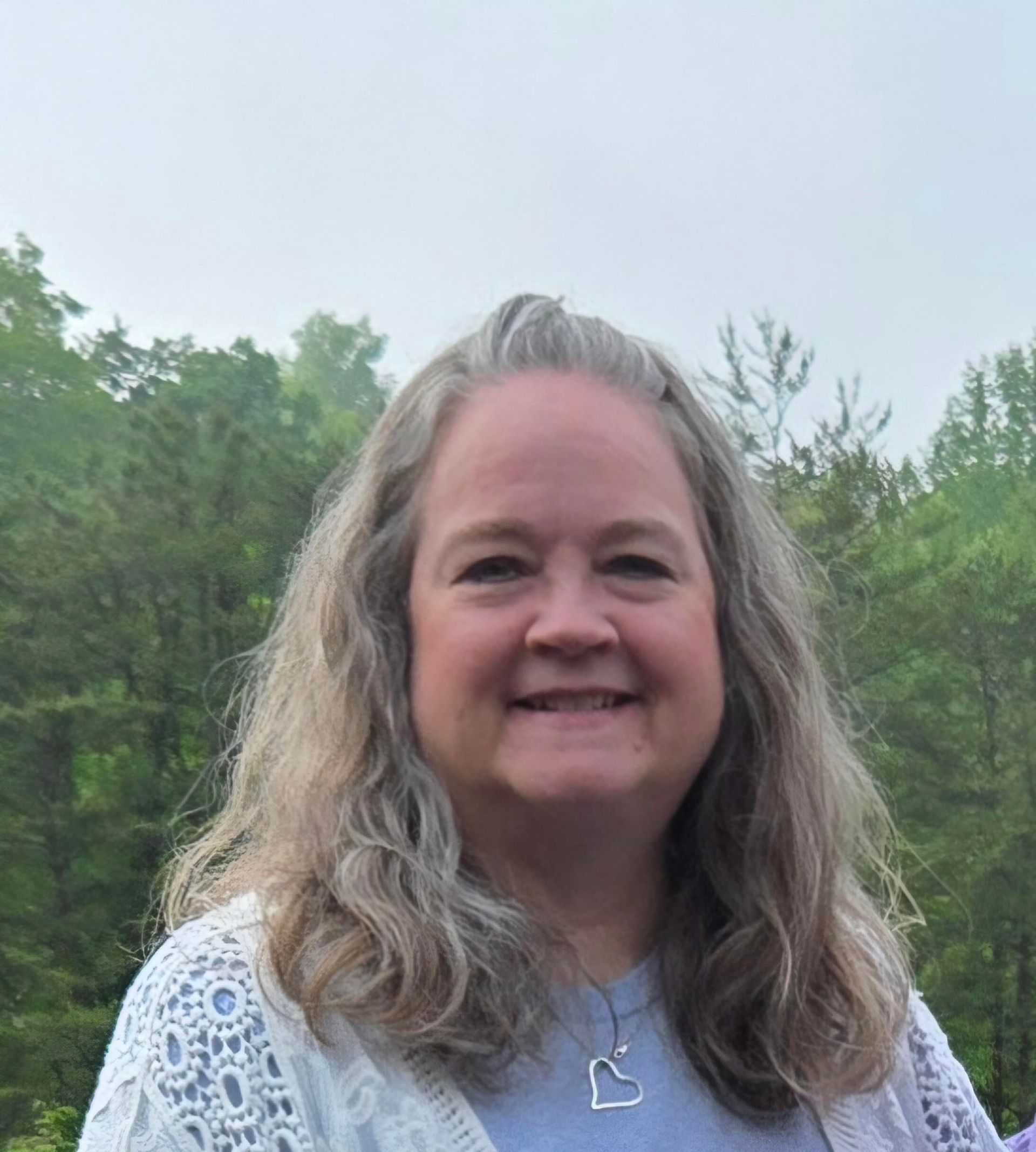Smiling person with long gray hair wearing a white lace top outdoors against trees