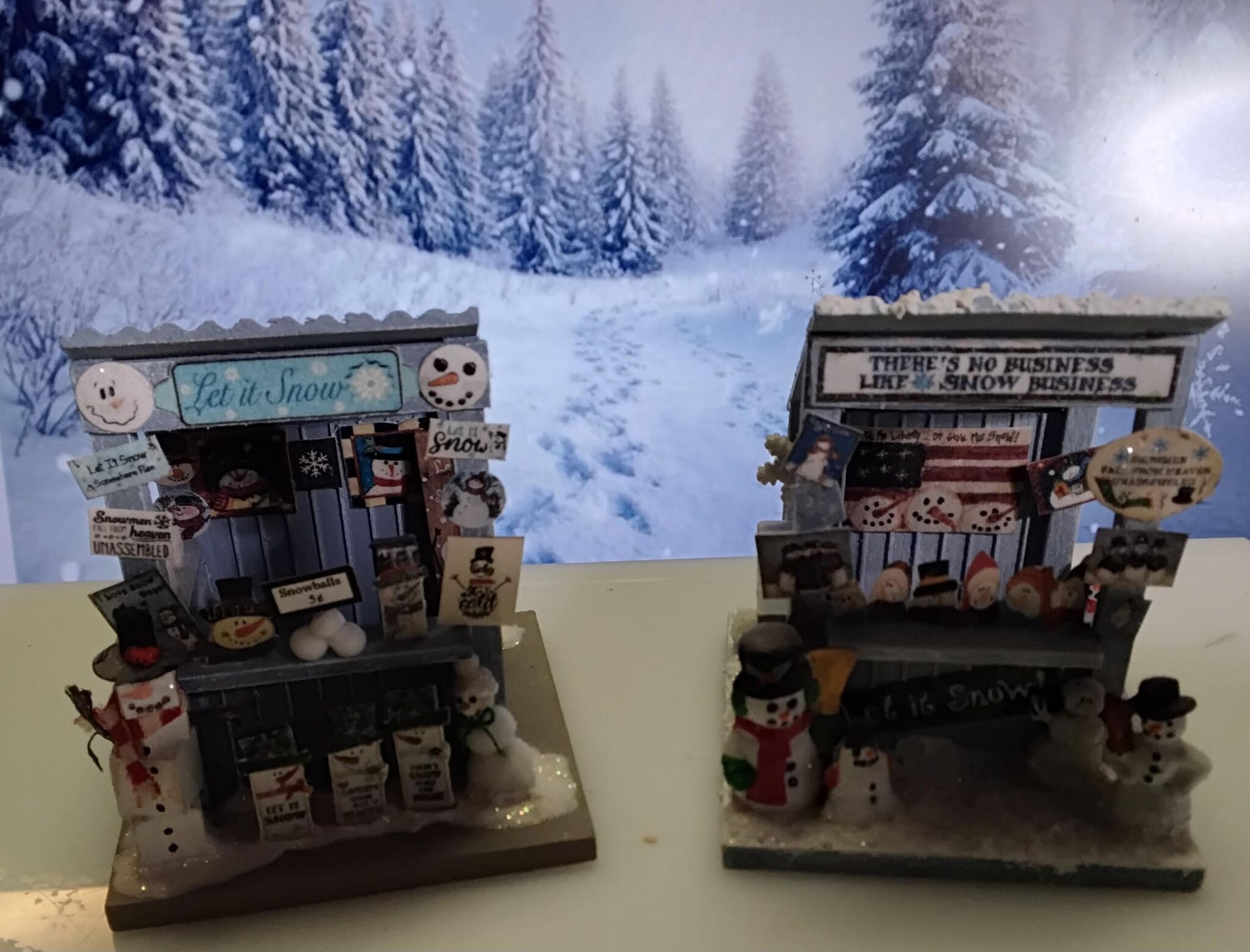 Two miniature holiday shop displays on a table before a snowy winter backdrop