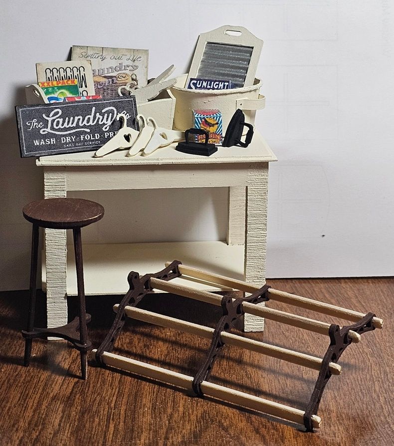 Miniature laundry room scene with a white counter, stool, and an overturned folding rack on a brown floor.
