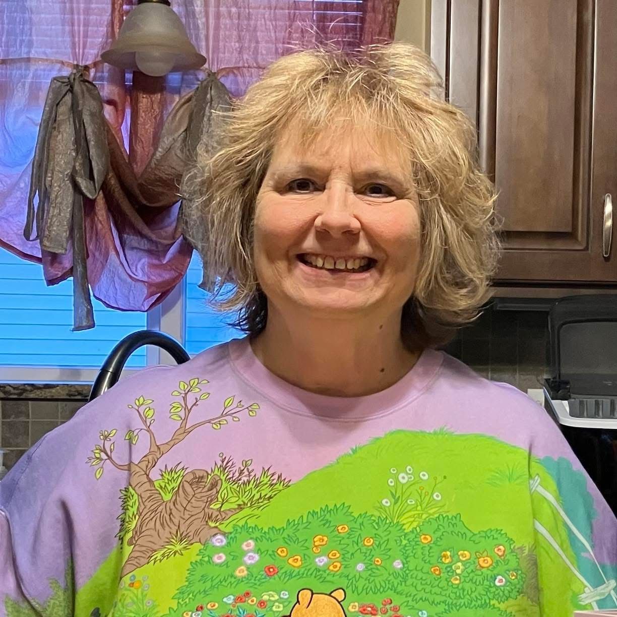 Smiling woman in a kitchen wearing a lavender floral shirt and standing by wooden cabinets