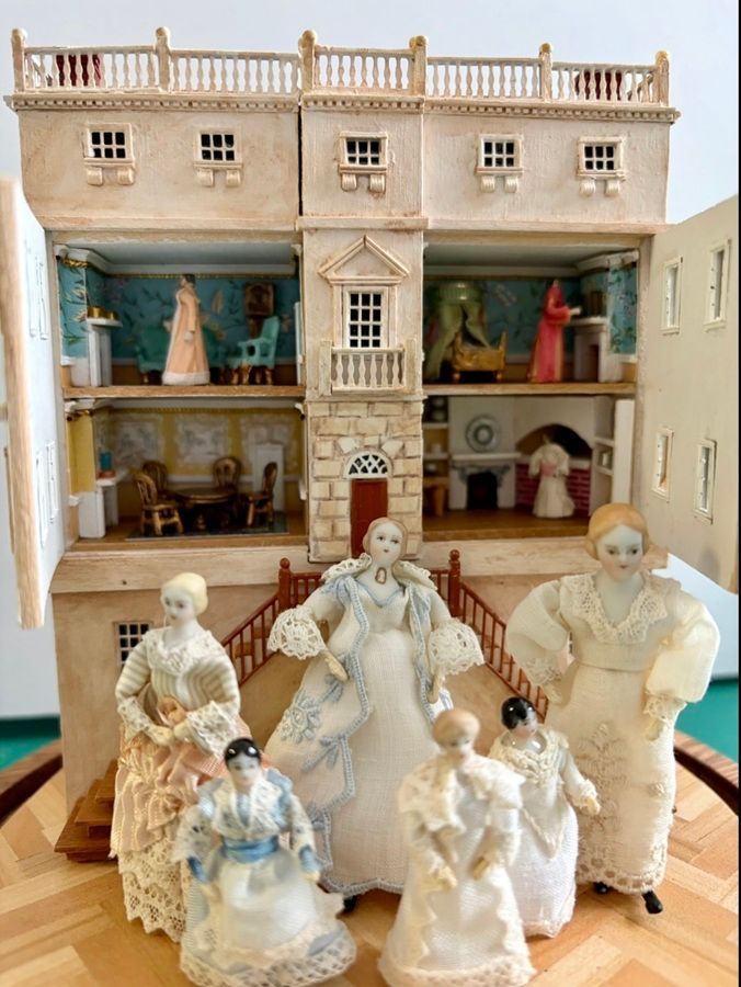 Miniature dollhouse facade with six porcelain figurines posed in front, in cream and teal tones.