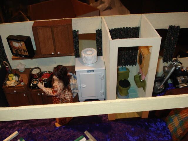Miniature dollhouse dining room with table, chairs, white cabinets, and two dolls in pastel clothing