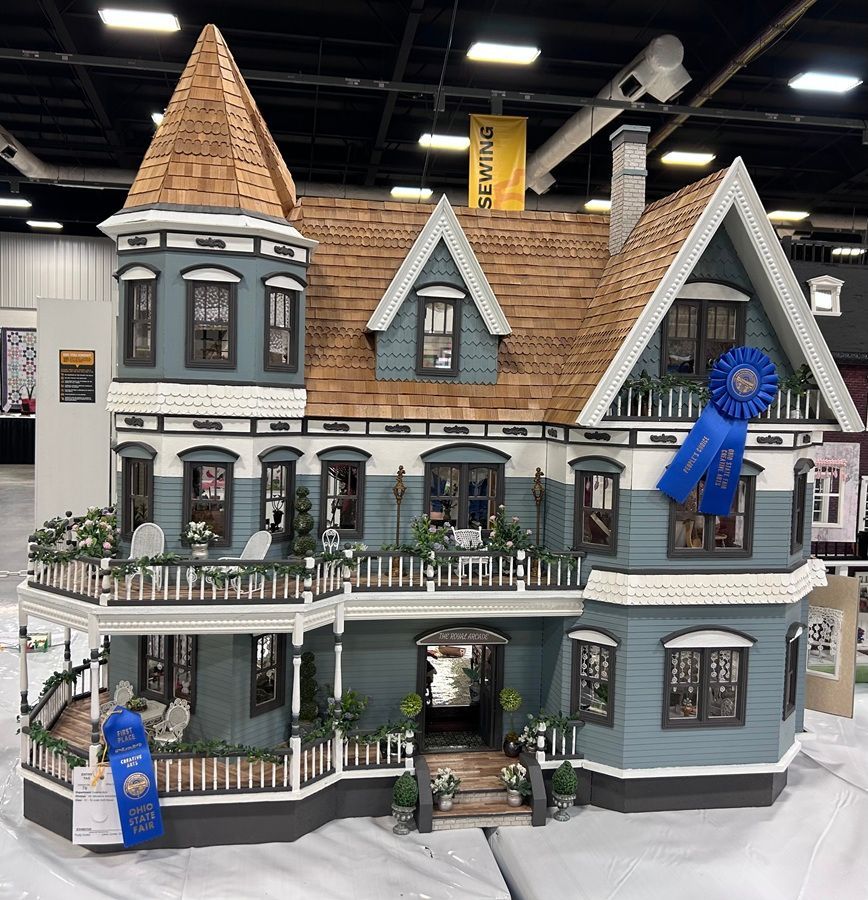 Victorian dollhouse with gray walls, brown shingle roof, blue ribbons, and a front porch on display indoors