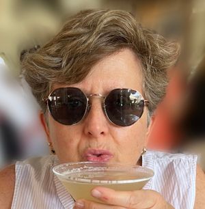 Person in sunglasses sipping a drink from a coupe glass indoors.