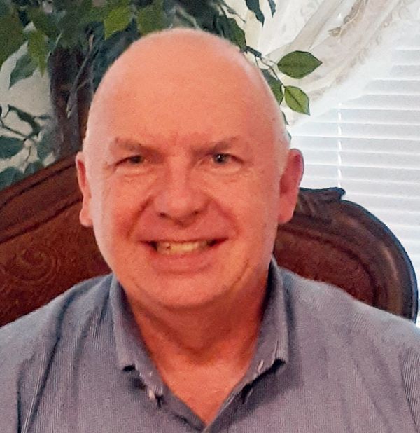Smiling bald man in a blue shirt seated indoors against a carved wooden chair and window blinds