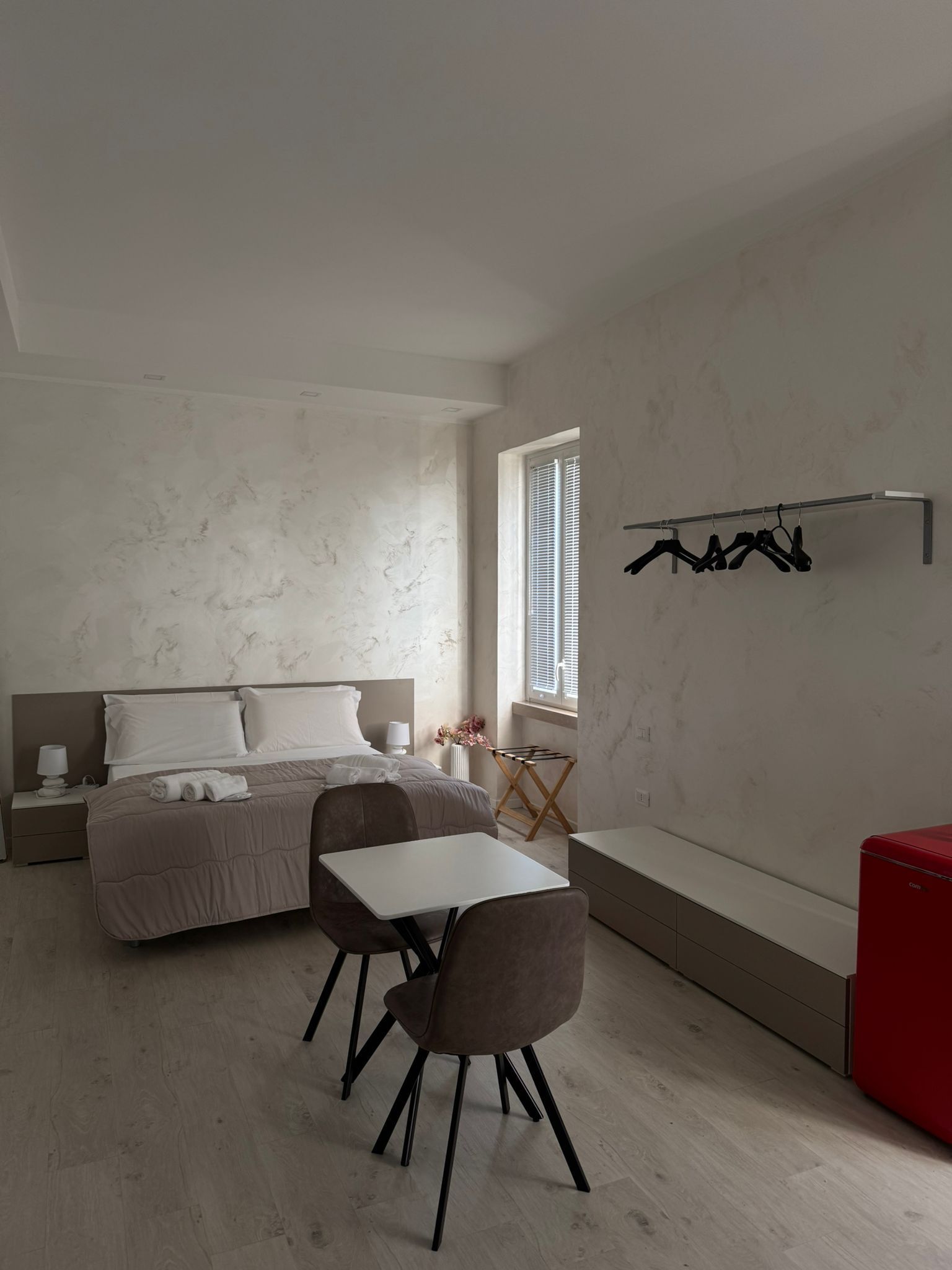 Camera da letto minimalista con letto, scrivania, sedia e appendiabiti a parete accanto a una finestra.
