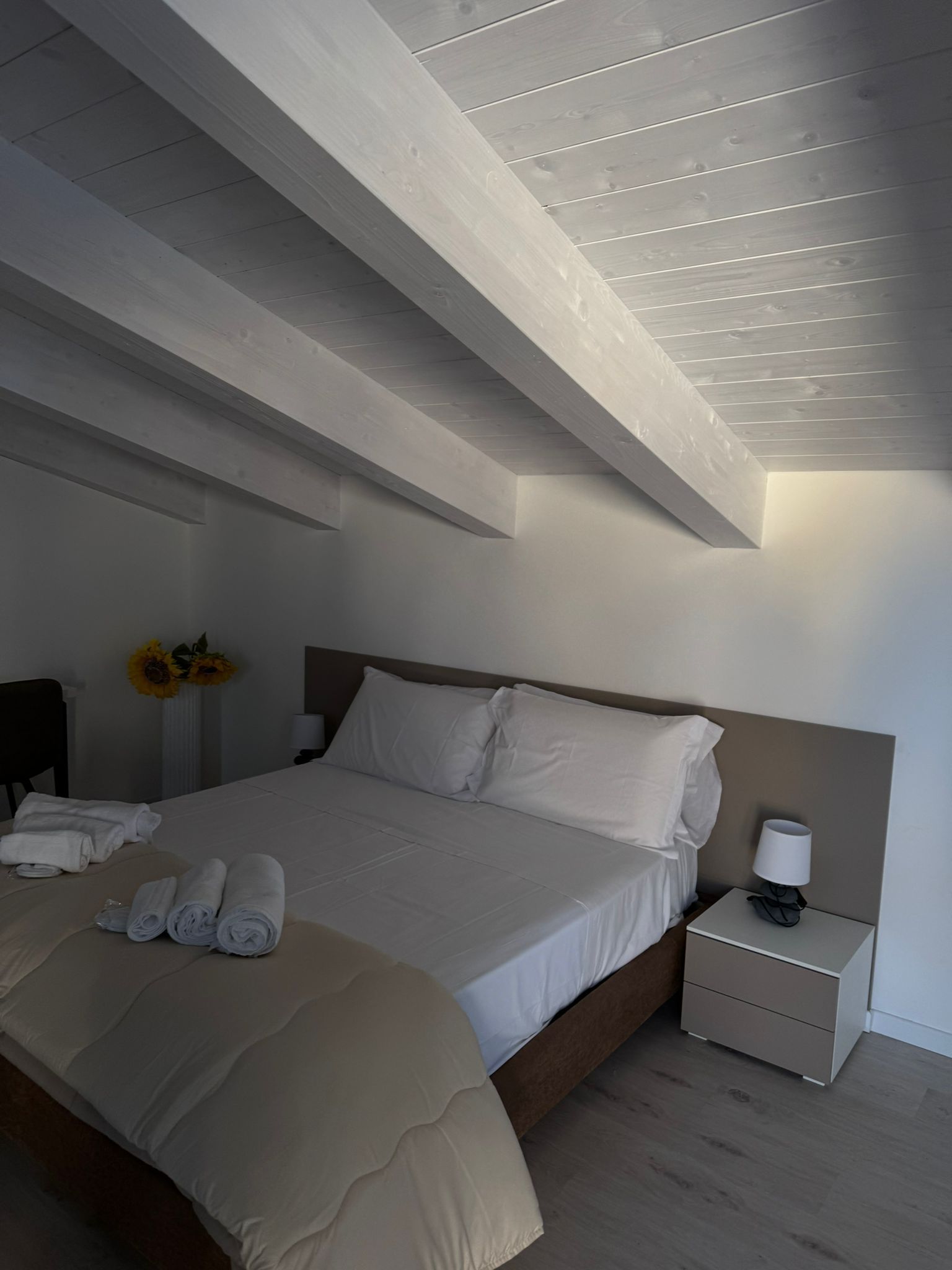 Piccola camera da letto bianca con soffitto spiovente, letto, comodini e morbida biancheria da letto grigia.