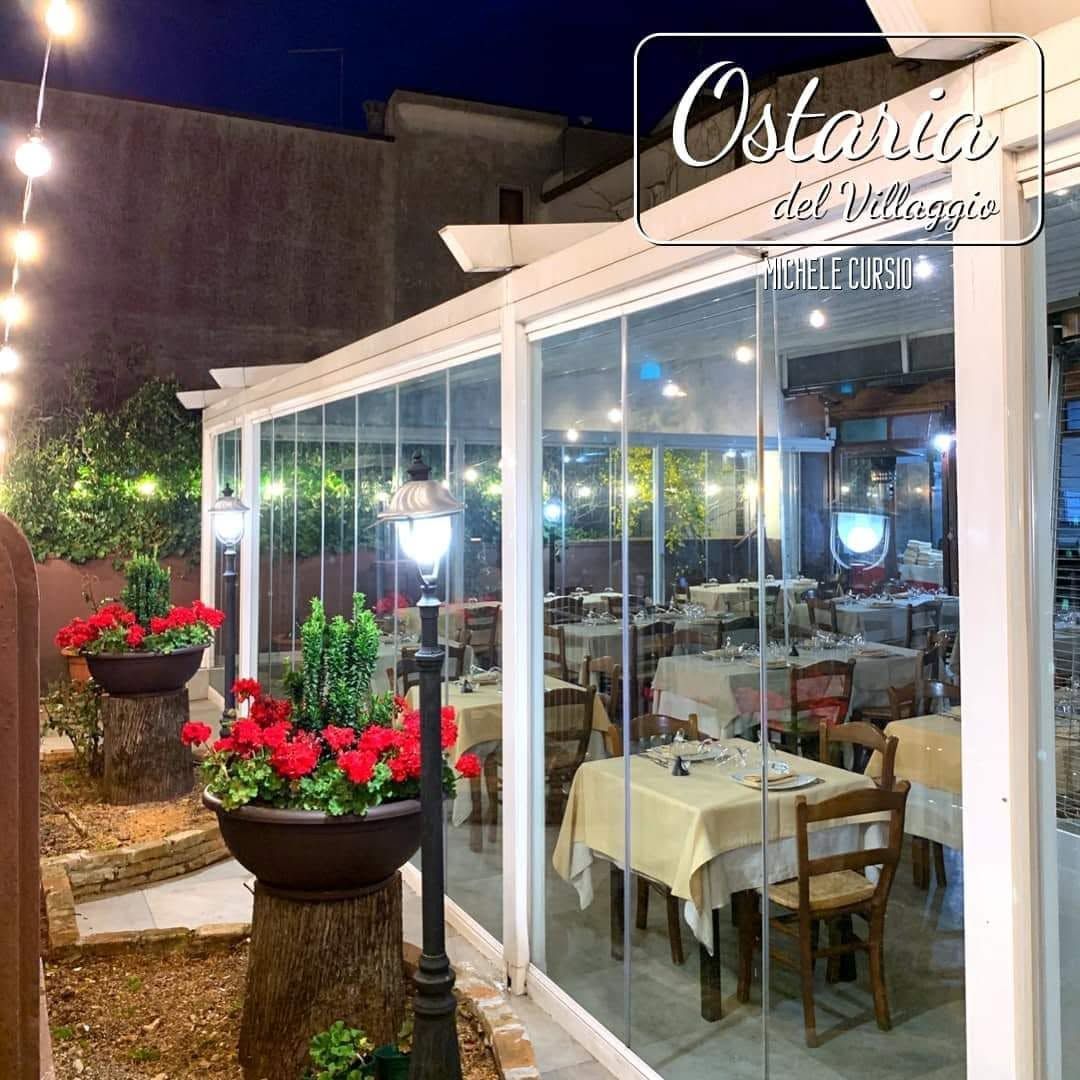 Terrazza esterna del ristorante di sera con pareti in vetro, tavoli bianchi e fiori rossi all'ingresso.