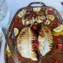 Pesce intero grigliato su un piatto, guarnito con spicchi di limone e pomodorini, servito con pinze e coltello.