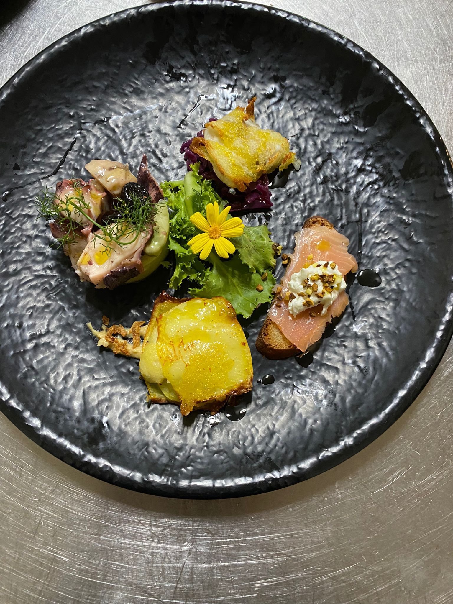 Quattro stuzzichini gourmet su un piatto nero, guarniti con fiori gialli ed erbe aromatiche.