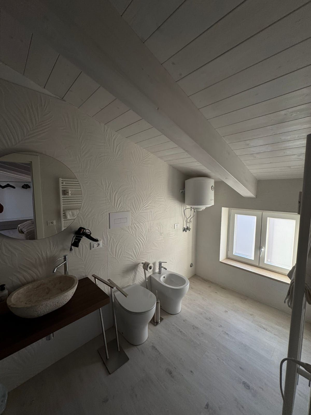Piccolo bagno bianco con lavandino, WC, doccia e due finestre sotto un soffitto inclinato.