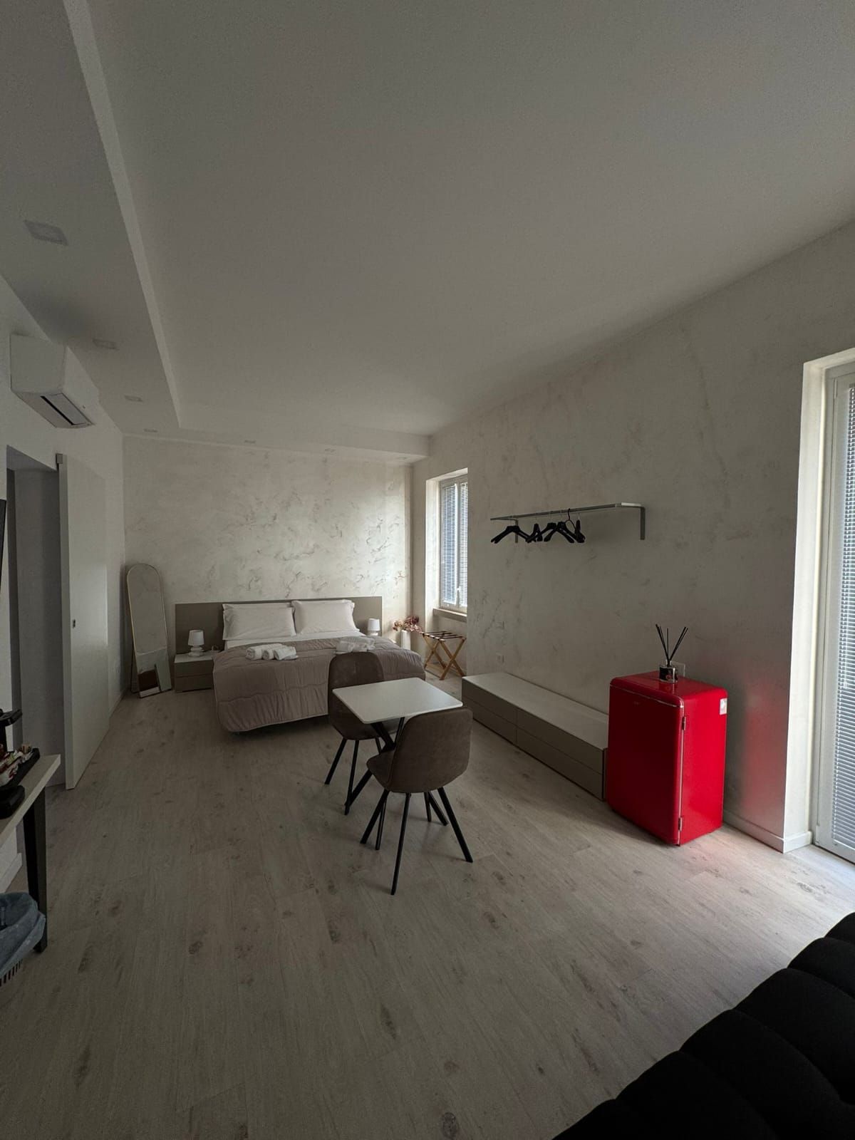 Camera da letto moderna con pareti bianche, letto, sedia, quadri e una valigia rossa vicino alla porta aperta.