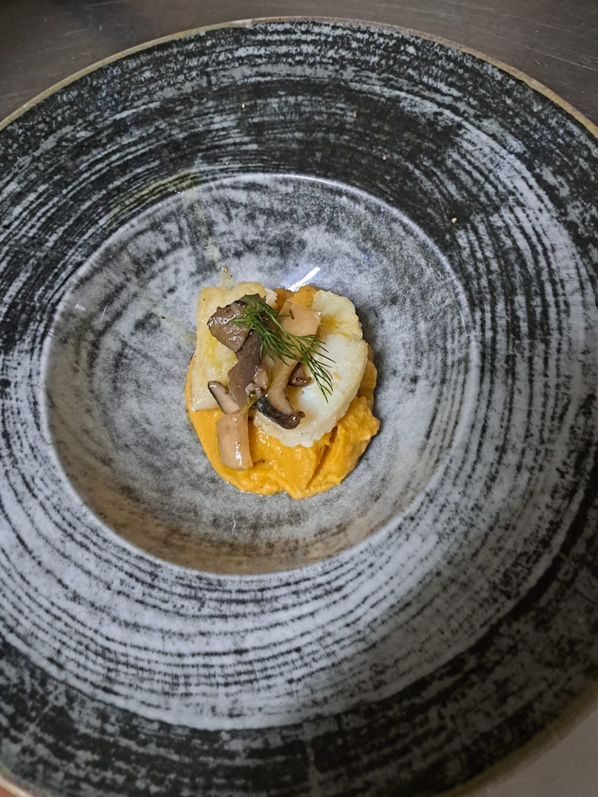 Piatto gourmet a base di funghi ed erbe aromatiche, servito su una purea arancione brillante in una ciotola scura dalla consistenza particolare.