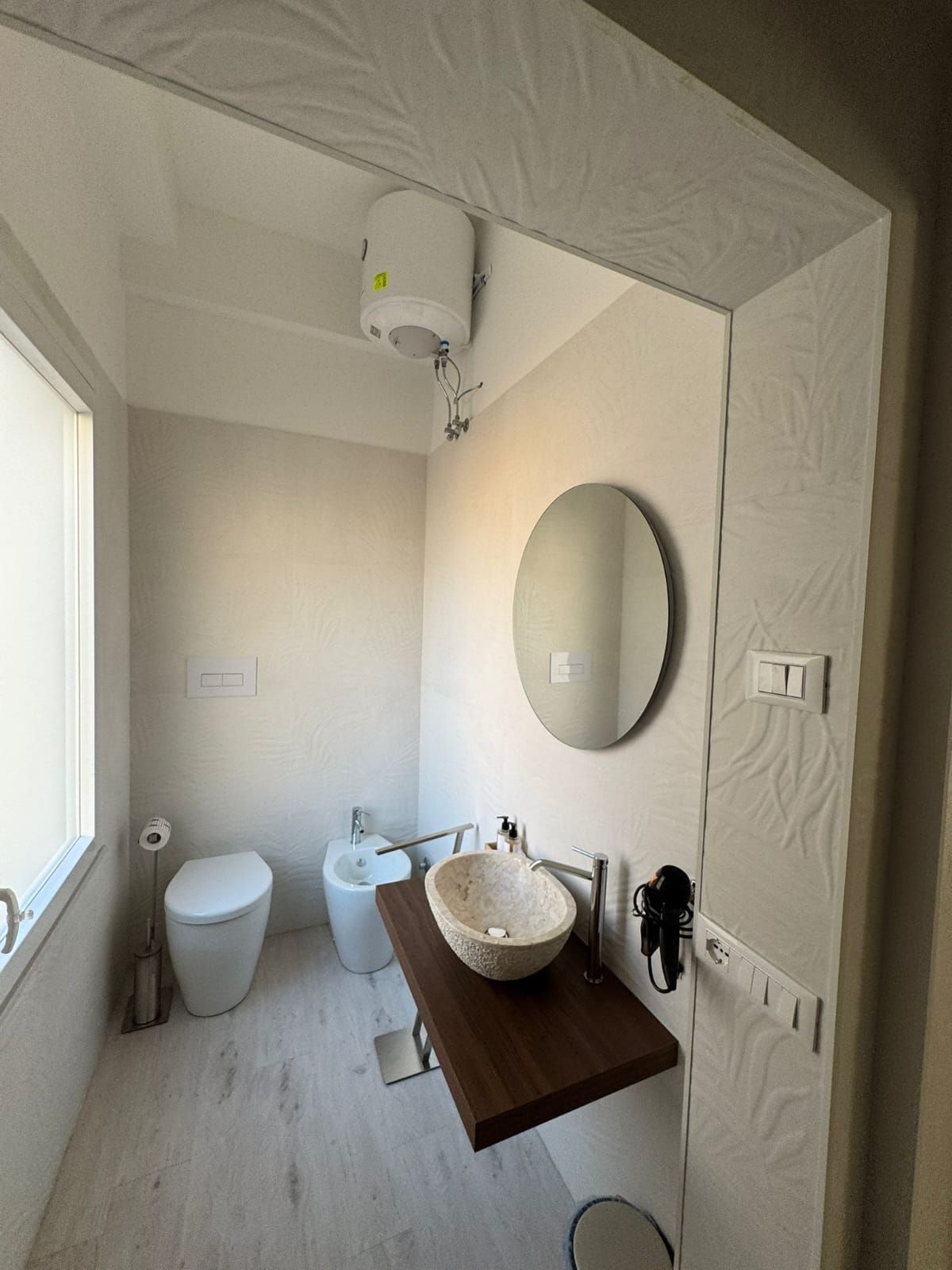 Piccolo bagno con WC, bidet, lavandino, specchio rotondo e luce a finestra.