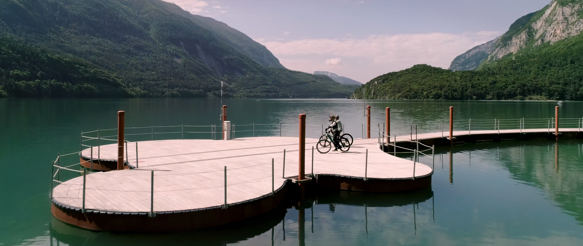 A lakeside dock with a bicycle on it, surrounded by water and mountains.