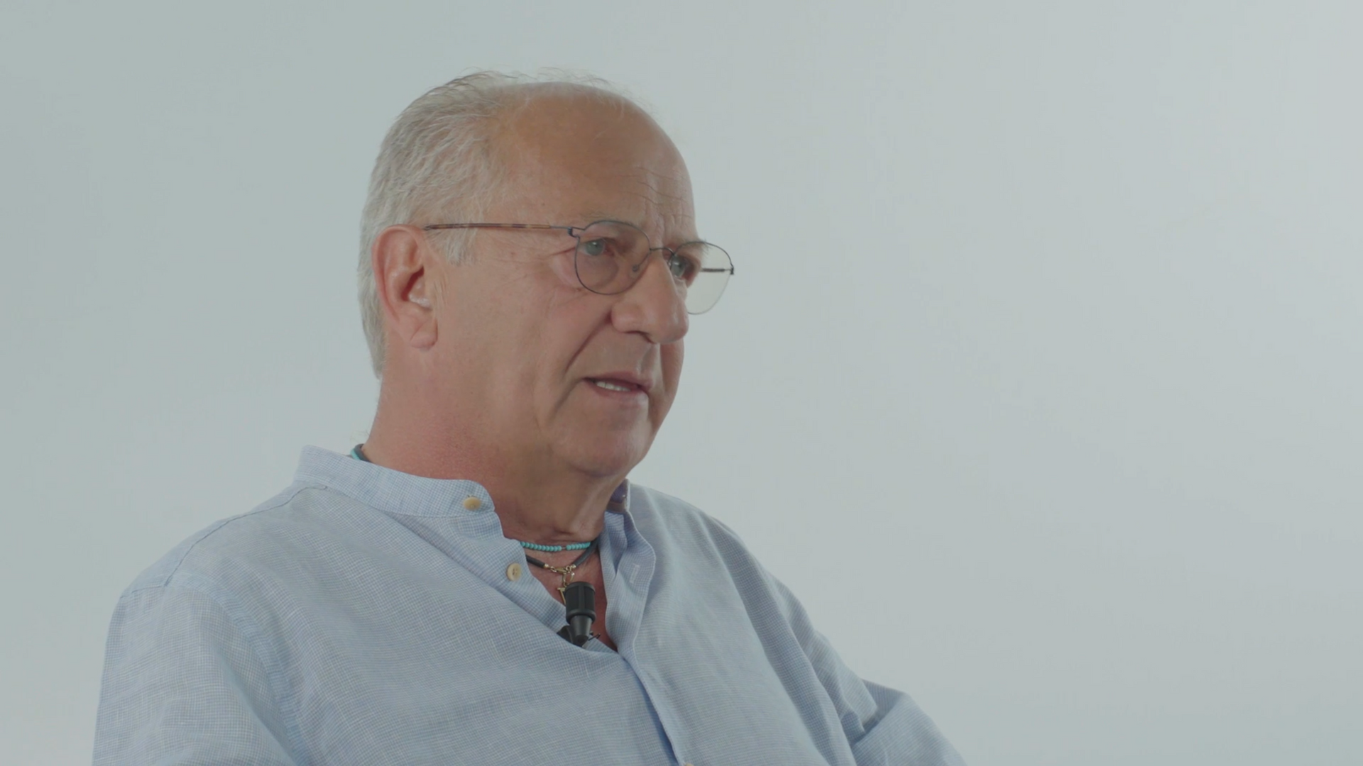 Man with glasses and a light blue shirt speaking, against a white backdrop.