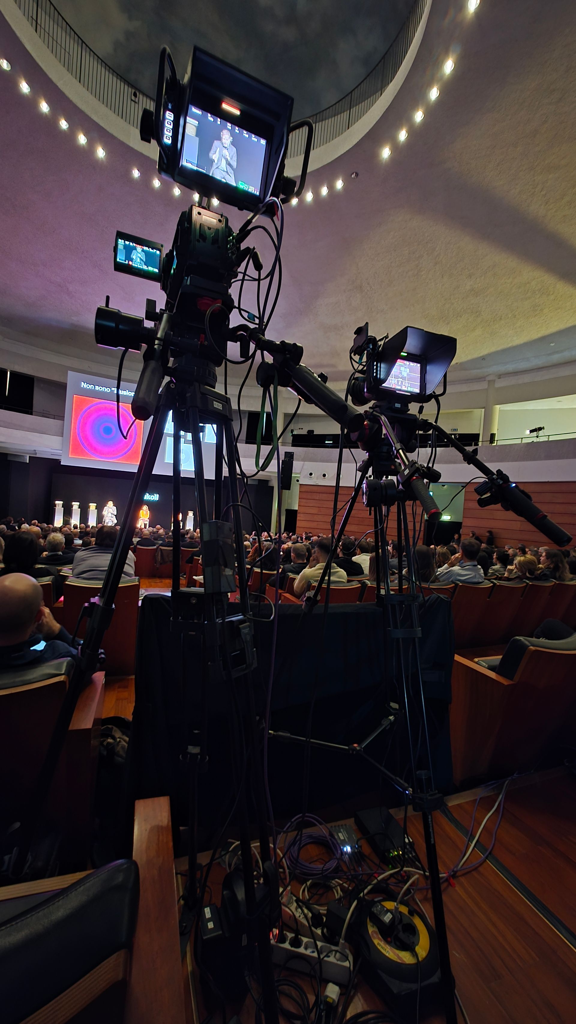 Due telecamere professionali su treppiedi all'interno di un grande auditorium, puntate su un evento con pubblico.