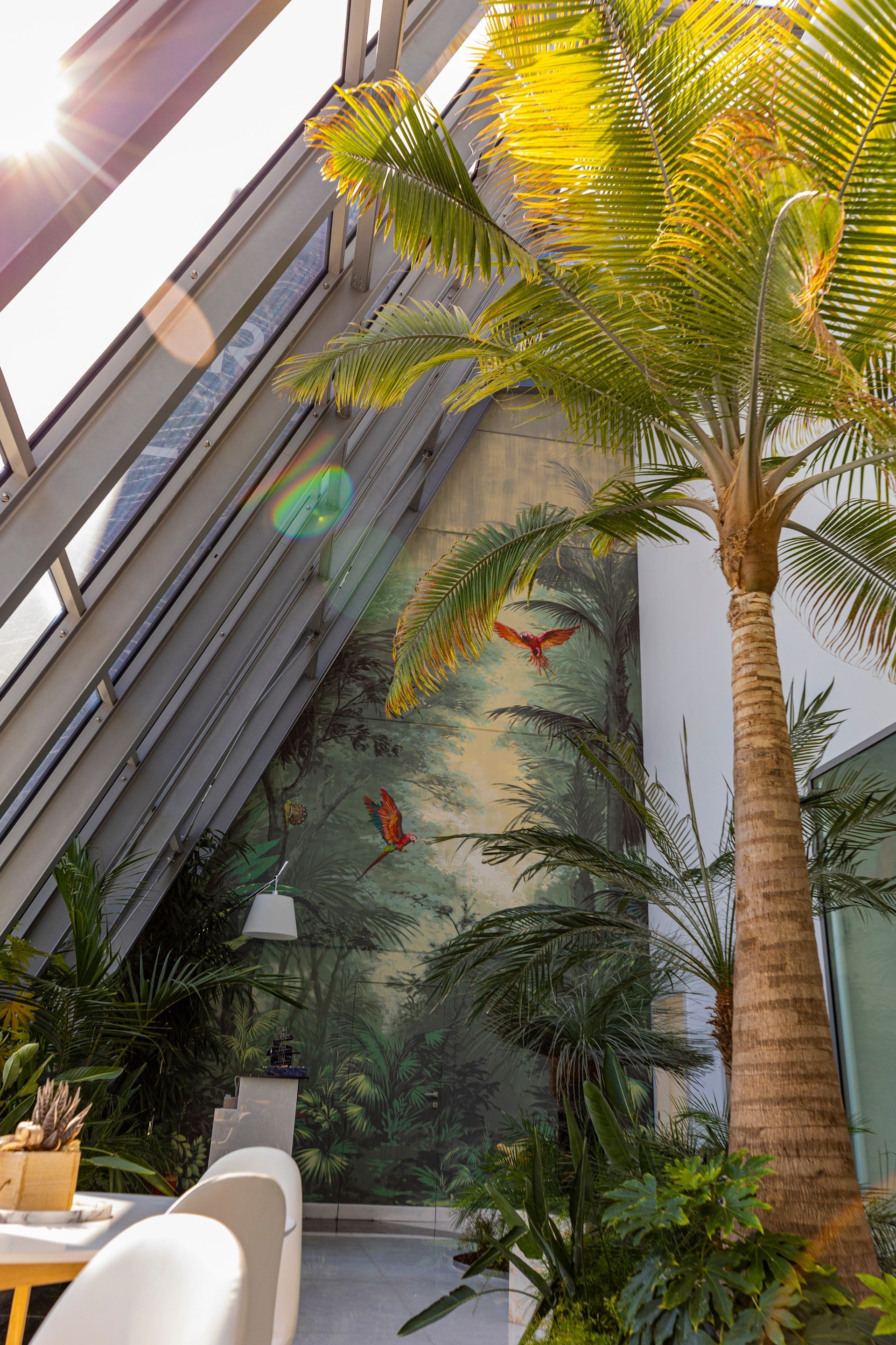 Una palma accanto a un murale tropicale, immersa nella luce del sole. Un interno moderno con soffitto in vetro.