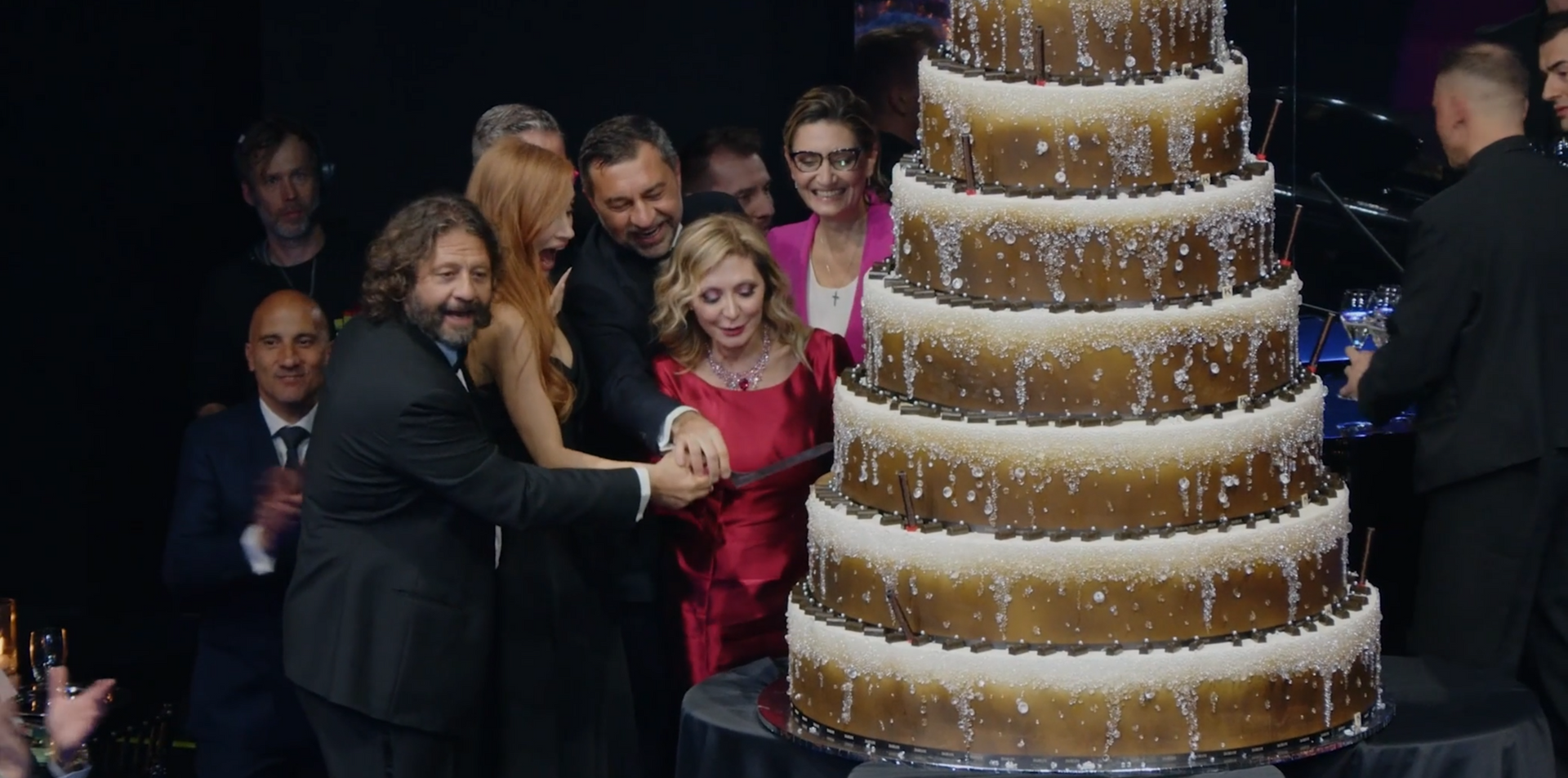 People cutting a large tiered cake on a stage; some smiling.