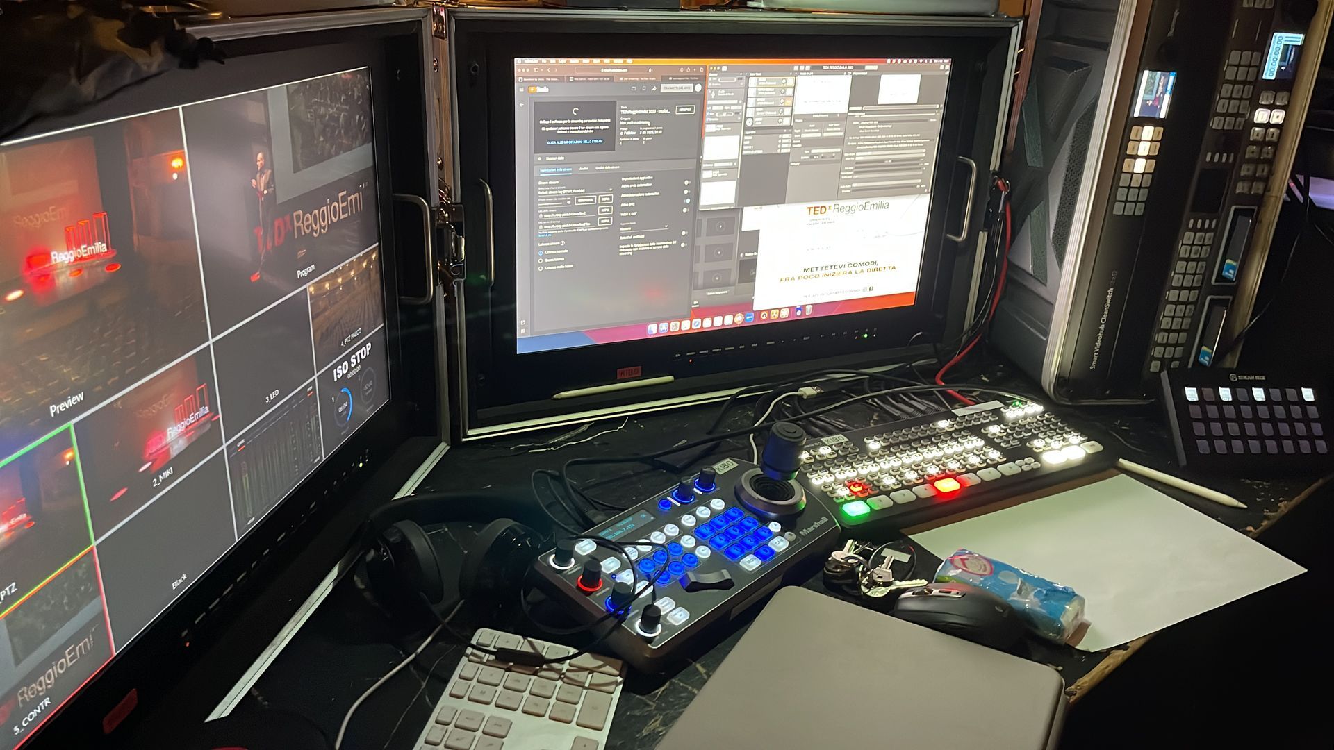 Pannello di controllo broadcast con due monitor che visualizzano i feed video, un joystick e una tastiera.