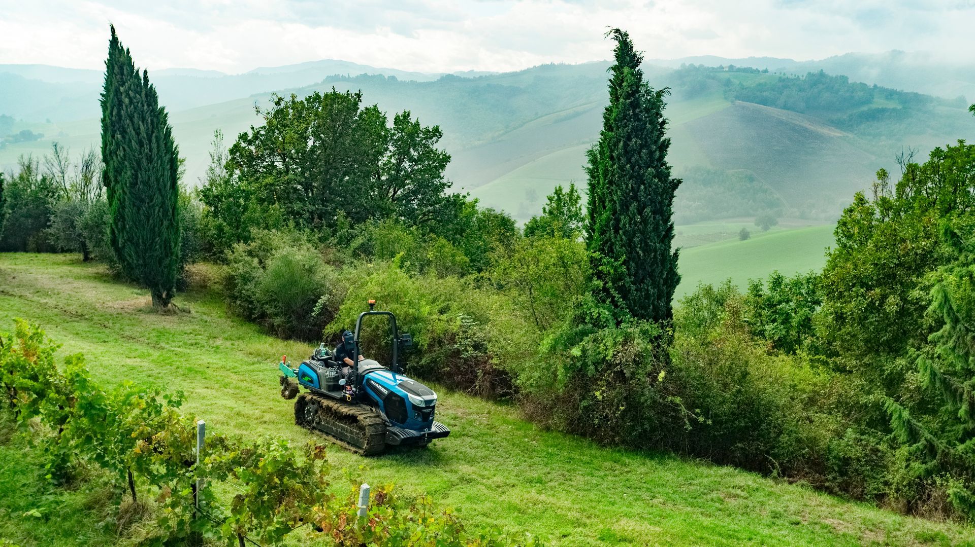 Tractor driving on a grassy hillside in Tuscany, Italy. Tall trees and rolling hills in the background.