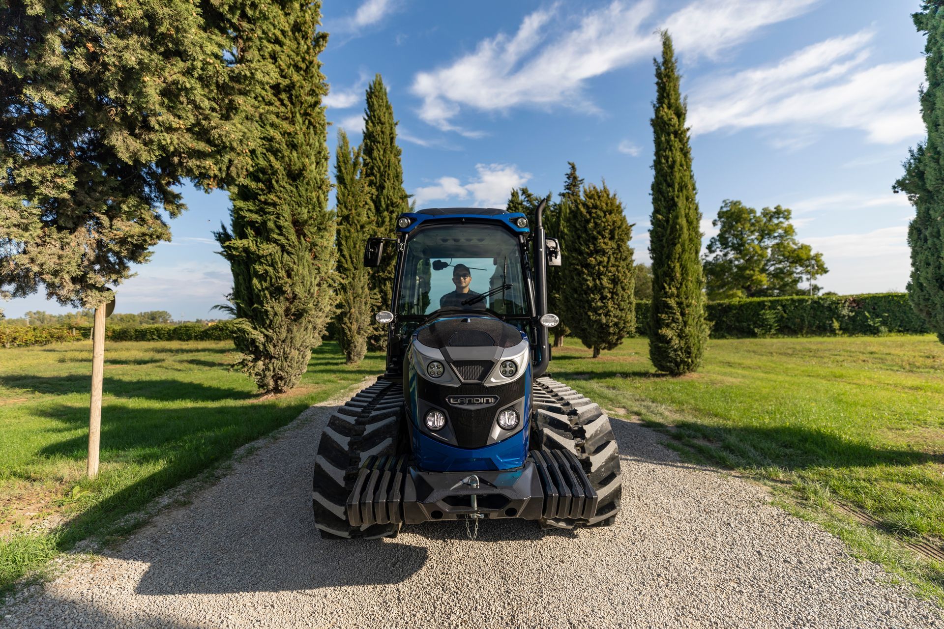 Blue tractor on a gravel path, flanked by trees; driver visible inside.