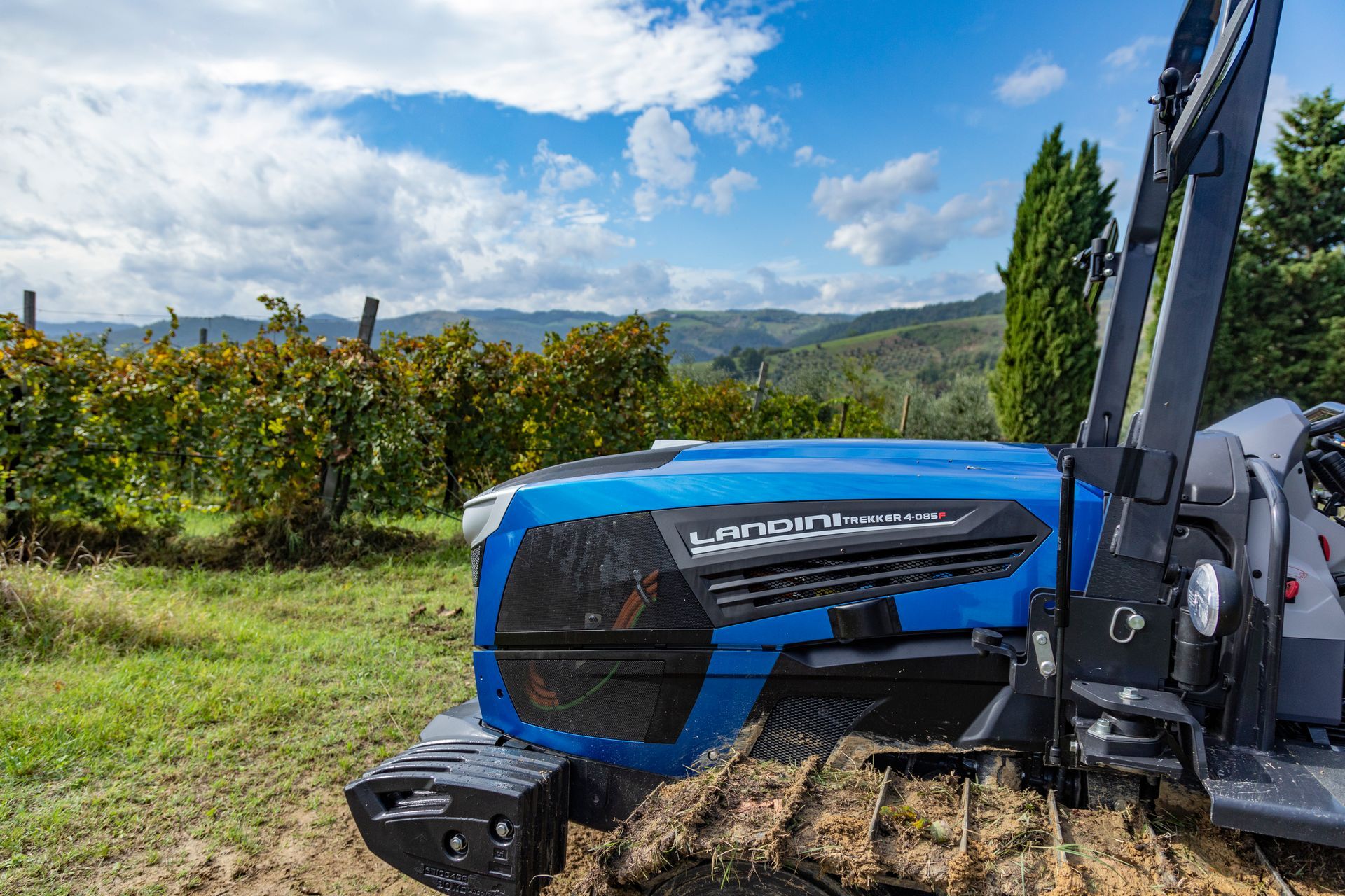 Blue tractor working in a vineyard, sunny day, with hills in the background.