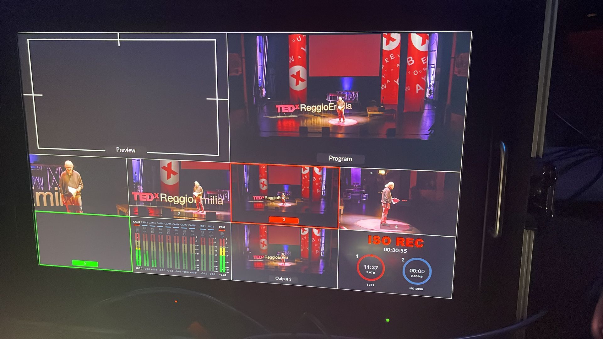 Schermo di produzione di un evento dal vivo che mostra diverse angolazioni di ripresa di un palco con un altoparlante e striscioni rossi.