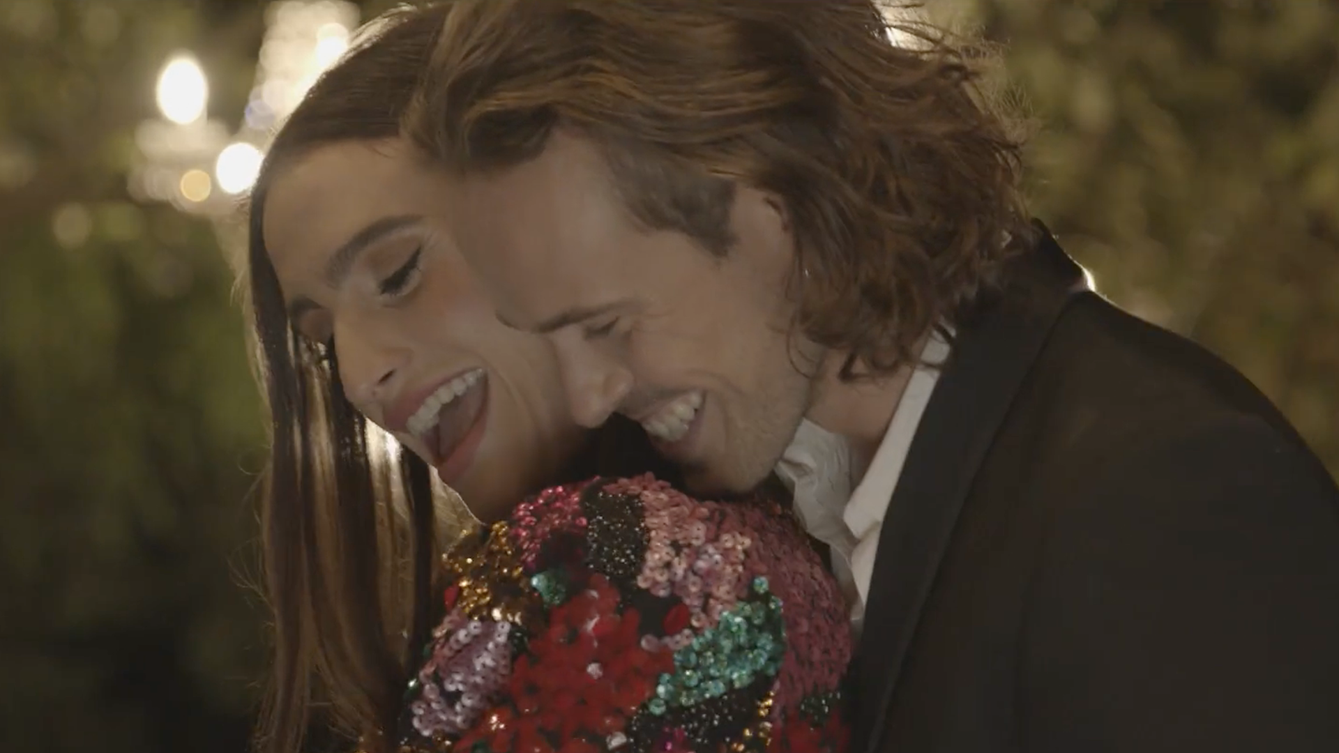 Smiling couple embracing, woman in sequined top, man in a suit, outdoor setting with lights.
