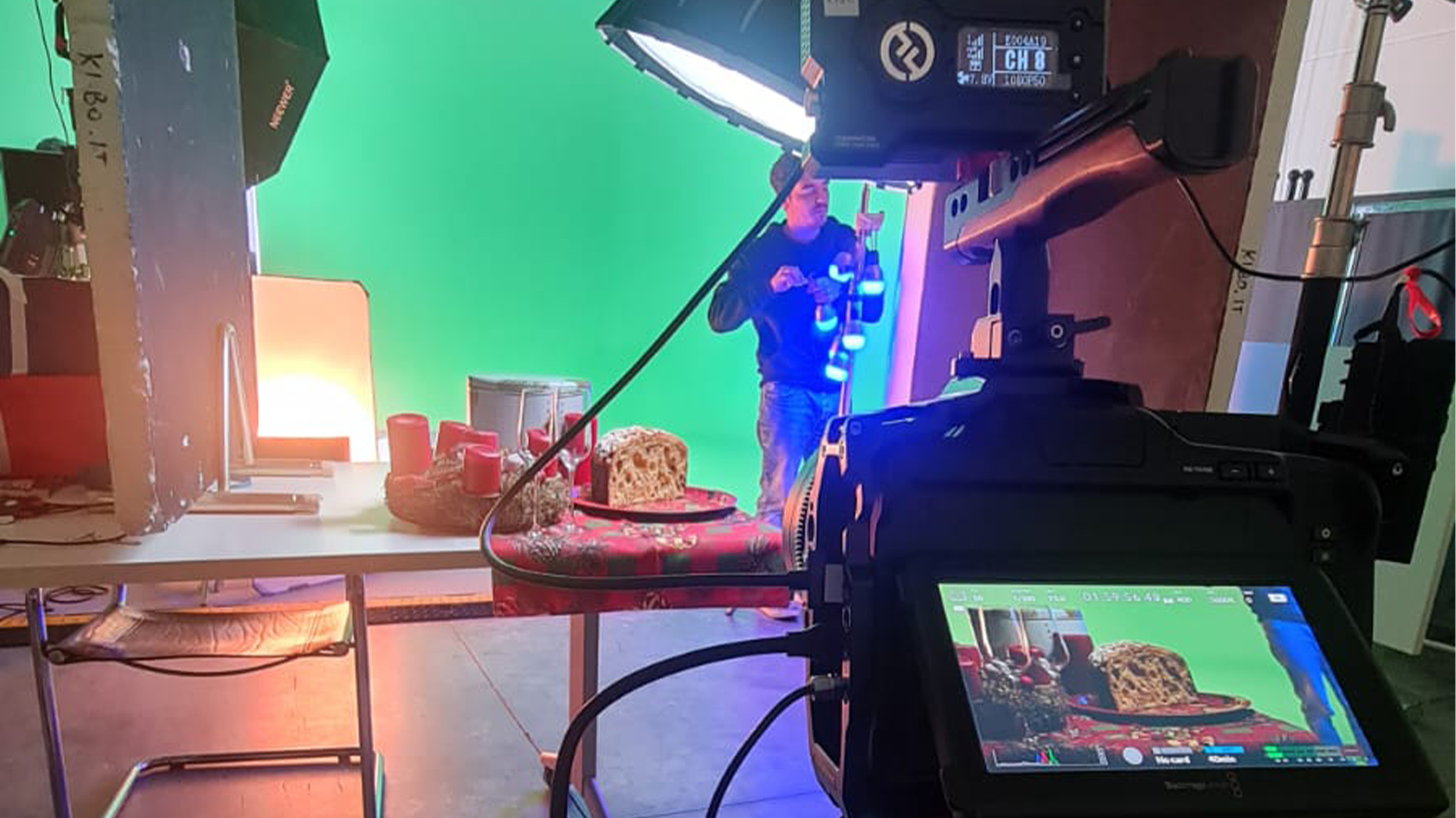Persona che filma una natura morta con una torta, davanti a uno schermo verde, luci e un monitor della telecamera.