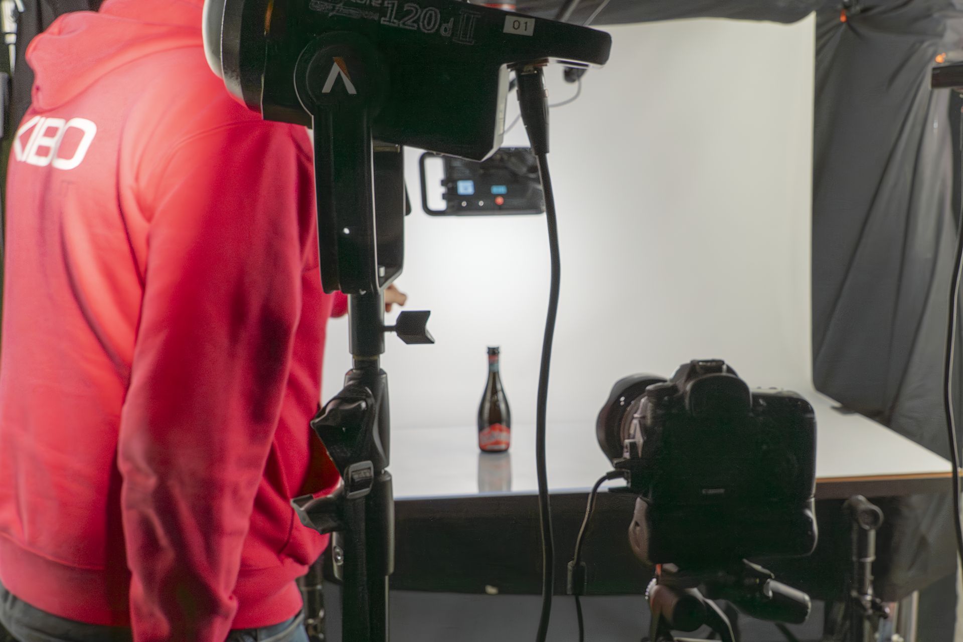 Persona con cappuccio rosso, bottiglia su un tavolo, illuminazione da studio, telecamera montata.