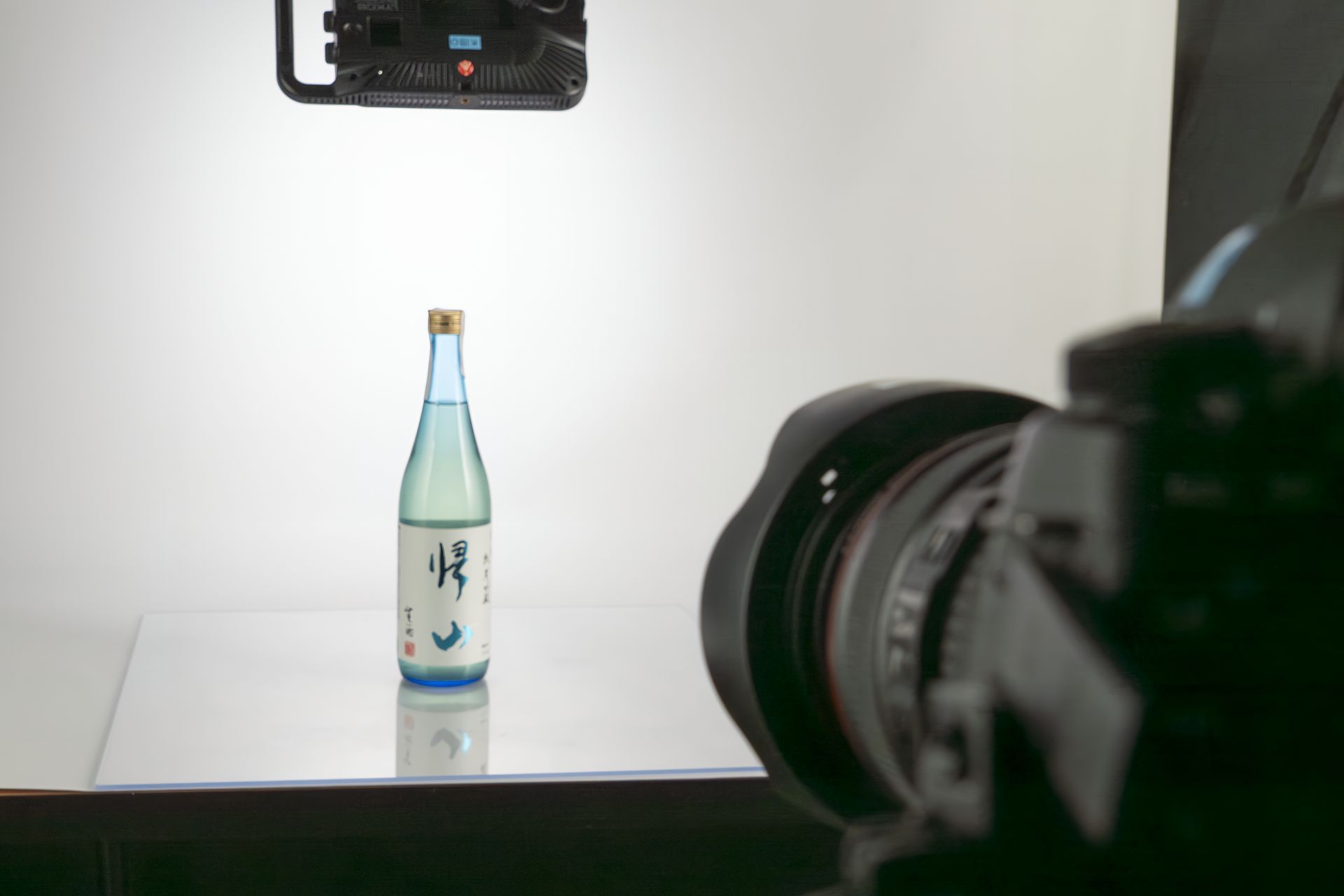 Bottiglia di sakè su una superficie riflettente, illuminata su uno sfondo bianco, con una macchina fotografica in primo piano.