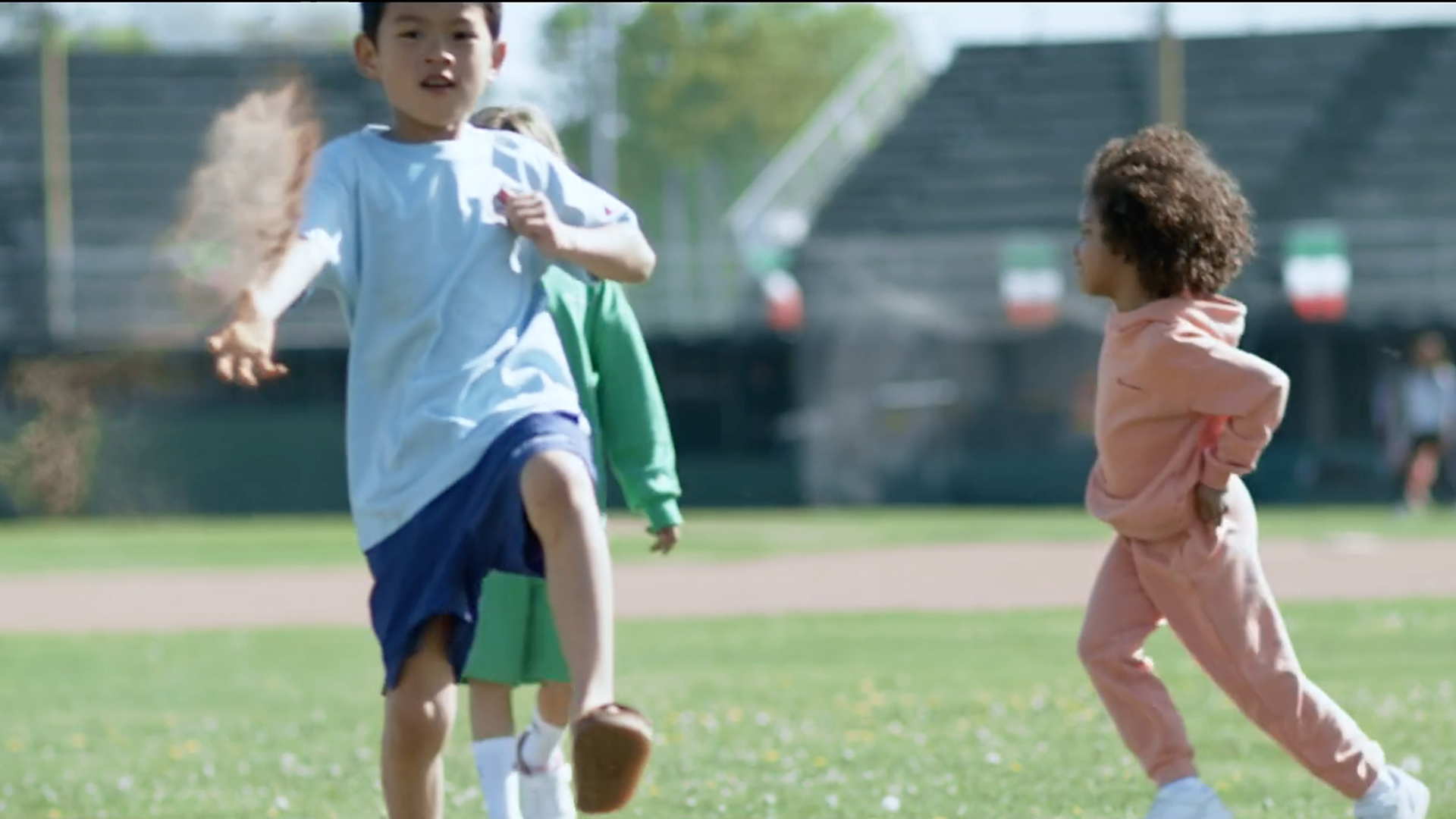 Bambini che giocano a rincorrersi su un campo erboso; uno lancia la terra, gli altri corrono; sullo sfondo lo stadio.
