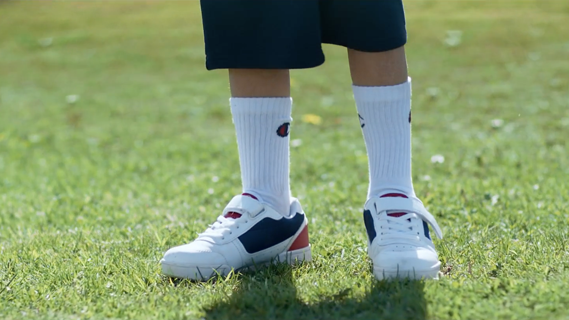 Gambe di una persona con scarpe da ginnastica bianche, calzini e pantaloncini blu su un prato verde.