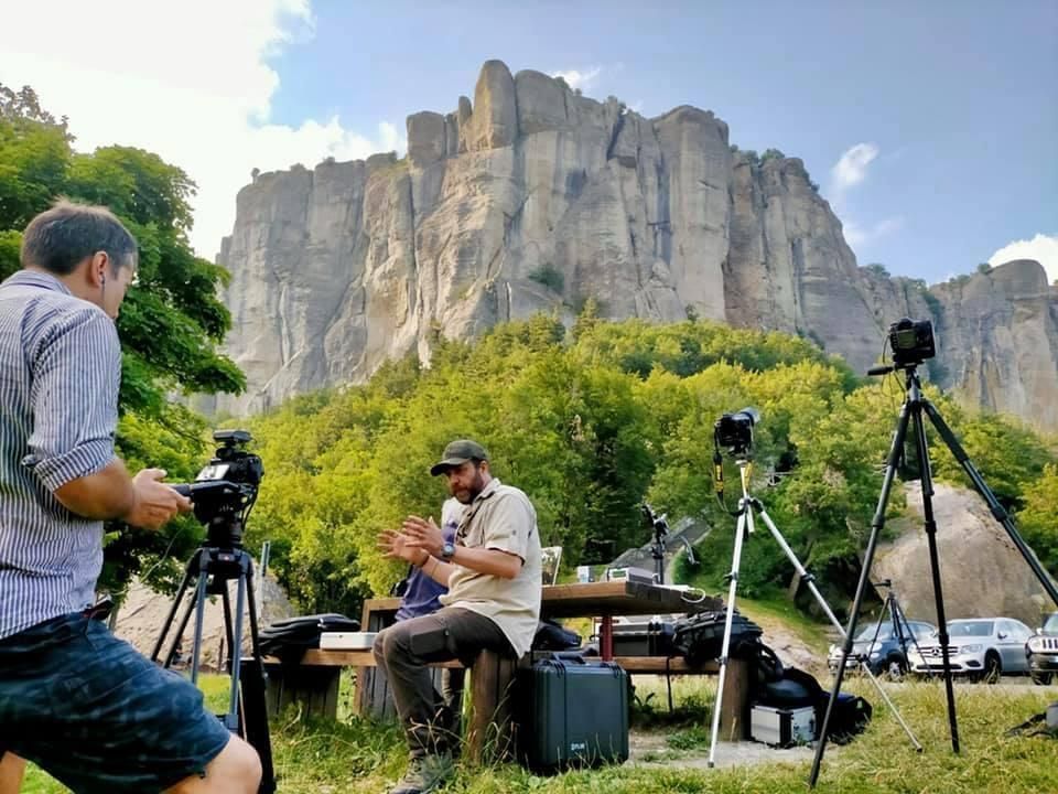 Two men filming outdoors with cameras on tripods, tall rock formation in background.