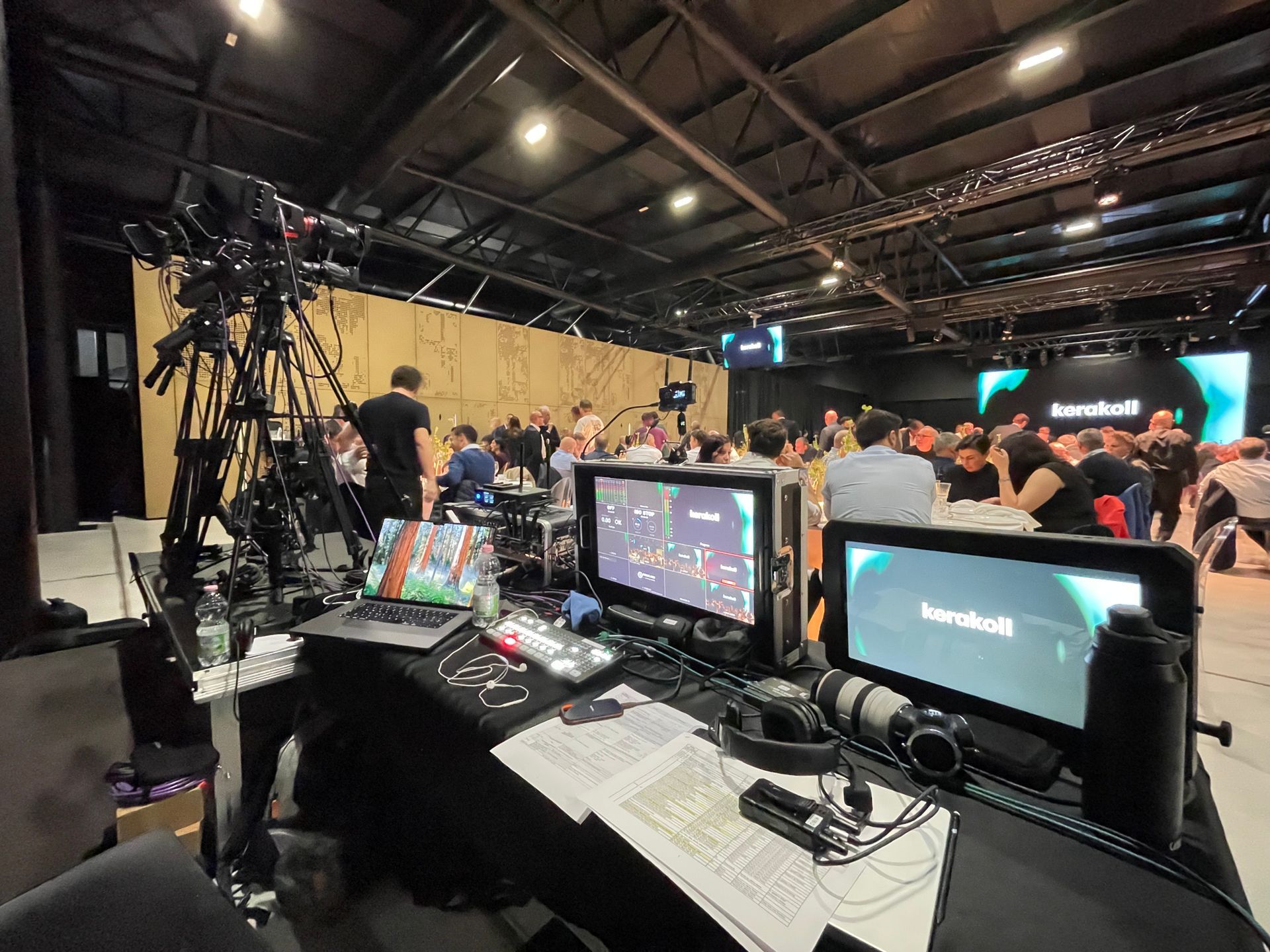 Un evento dal vivo in uno studio con telecamere, monitor e un pubblico di fronte a uno sfondo di legno.