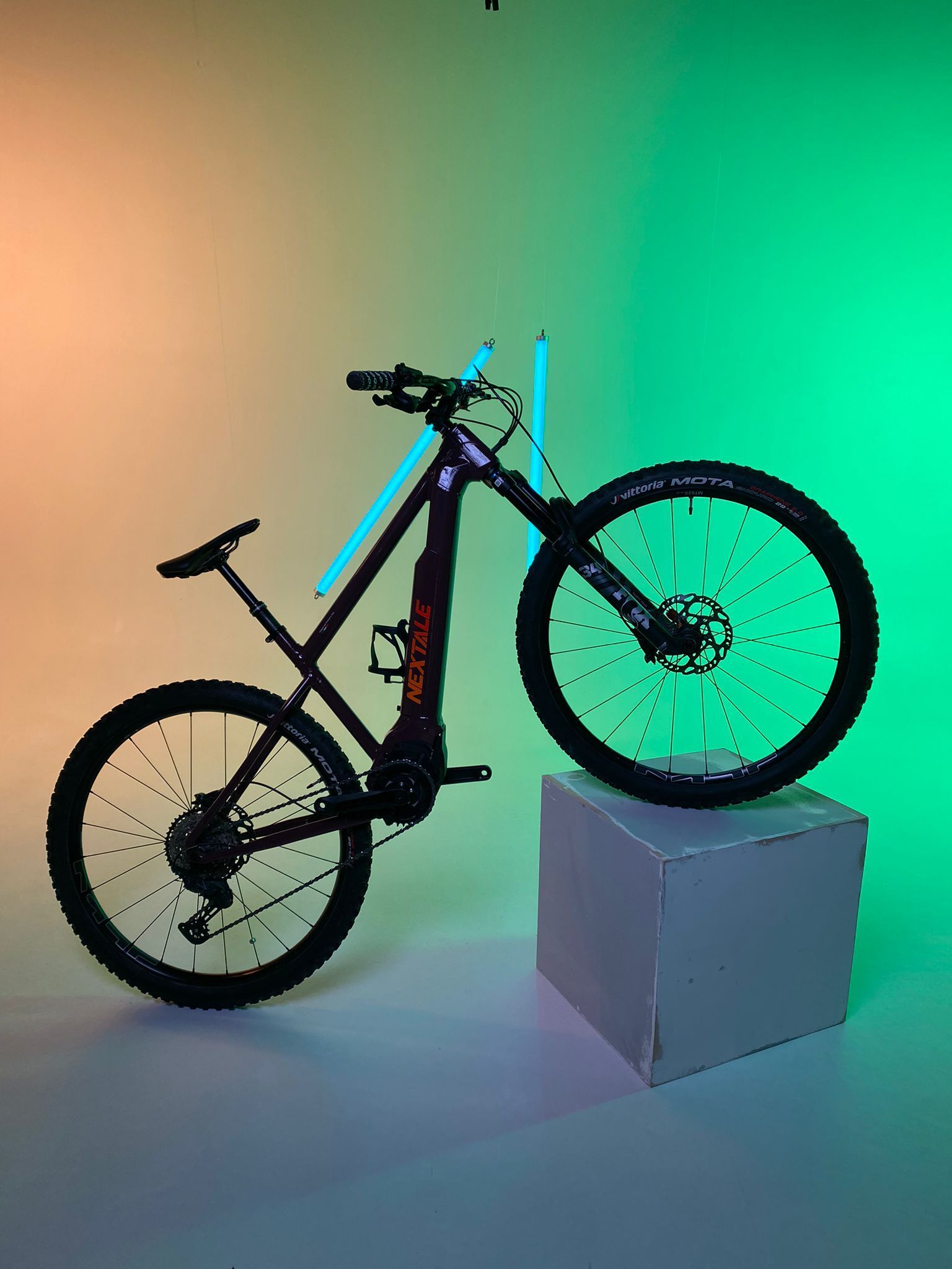 Black electric mountain bike on a white cube, with green and orange background lighting.