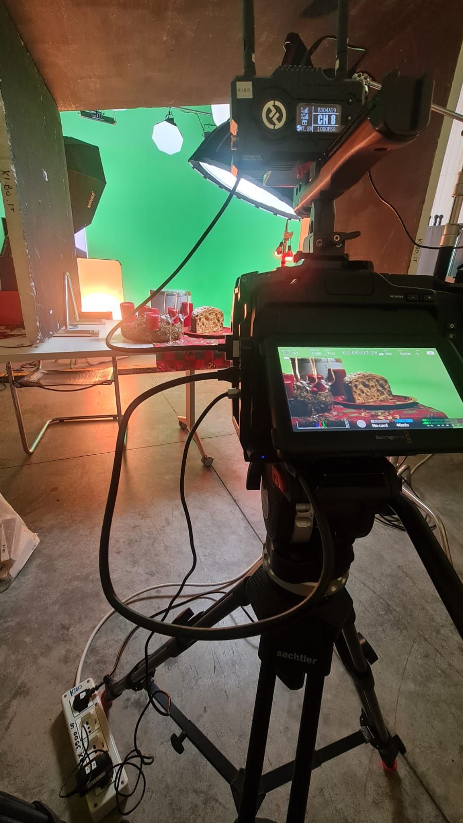 Un set cinematografico: una telecamera su un treppiede, uno sfondo verde, del cibo su un tavolo e delle luci.