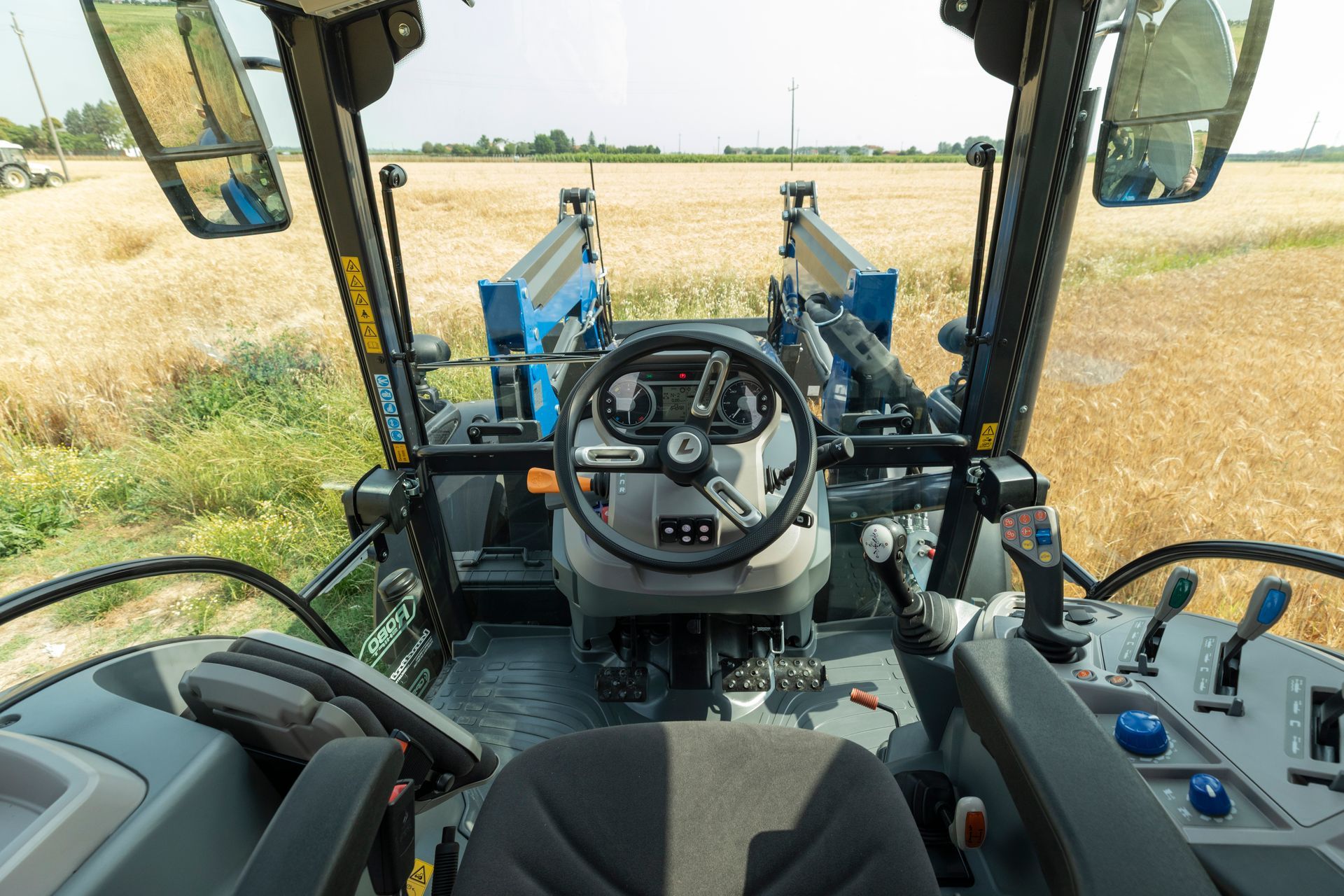 Vista interna della cabina di un trattore con volante, comandi e vista di un campo.